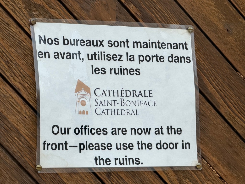 A sign reading (in French and English): Our offices are now at the front-- please use the door in the ruins.